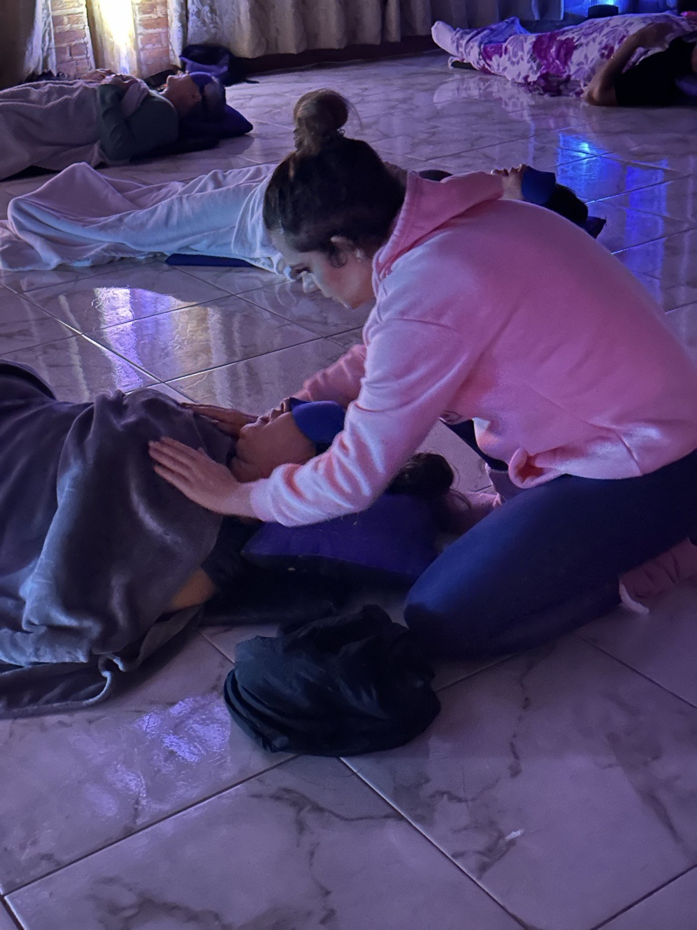 Facilitadora com as mãos sobre uma participante deitada, em luz roxa